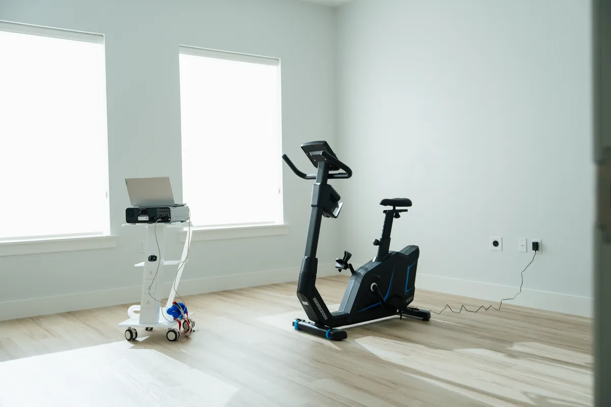 A stationary exercise bike and a wheeled equipment cart with a laptop in a bright, open room at Highland Longevity
